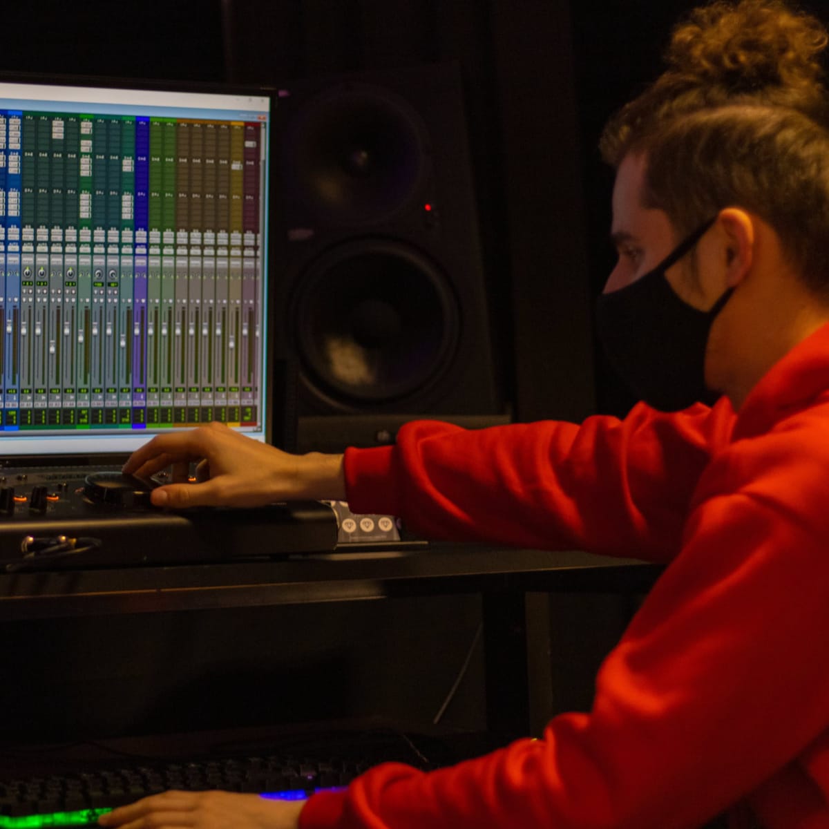 A person in a red sweatshirt works on a sound mixing software on a computer screen while another figure stands nearby.