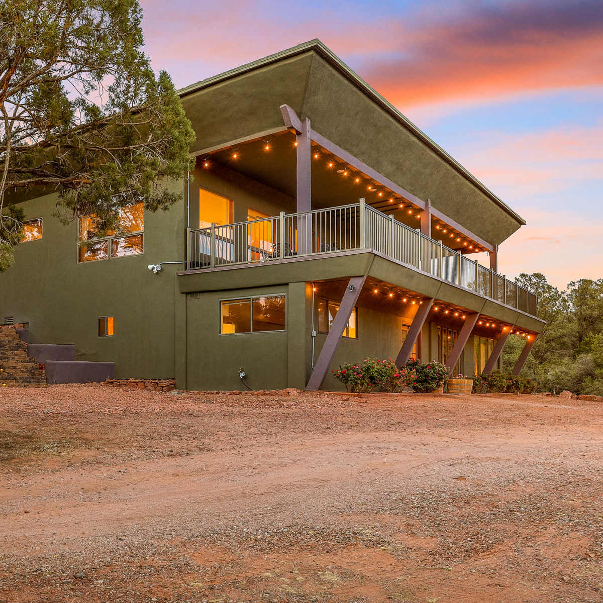 A modern two-story green house with a wraparound deck and string lights is situated on a gravel driveway surrounded by trees.