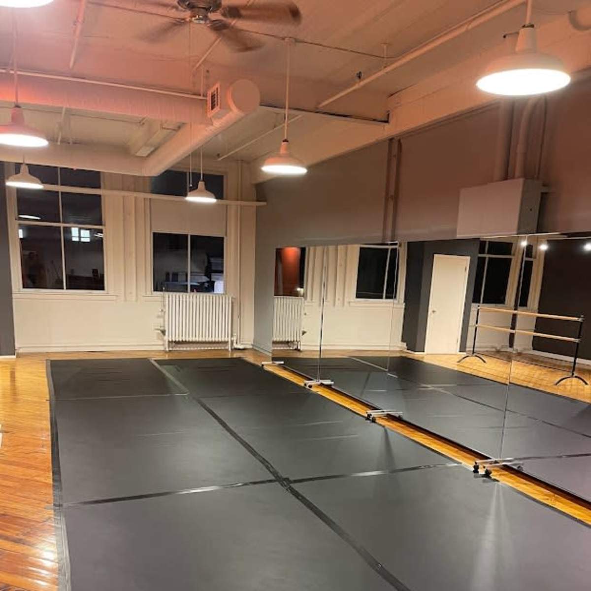 A dance studio featuring a black rubber floor, large mirrors on one wall, and overhead lighting.