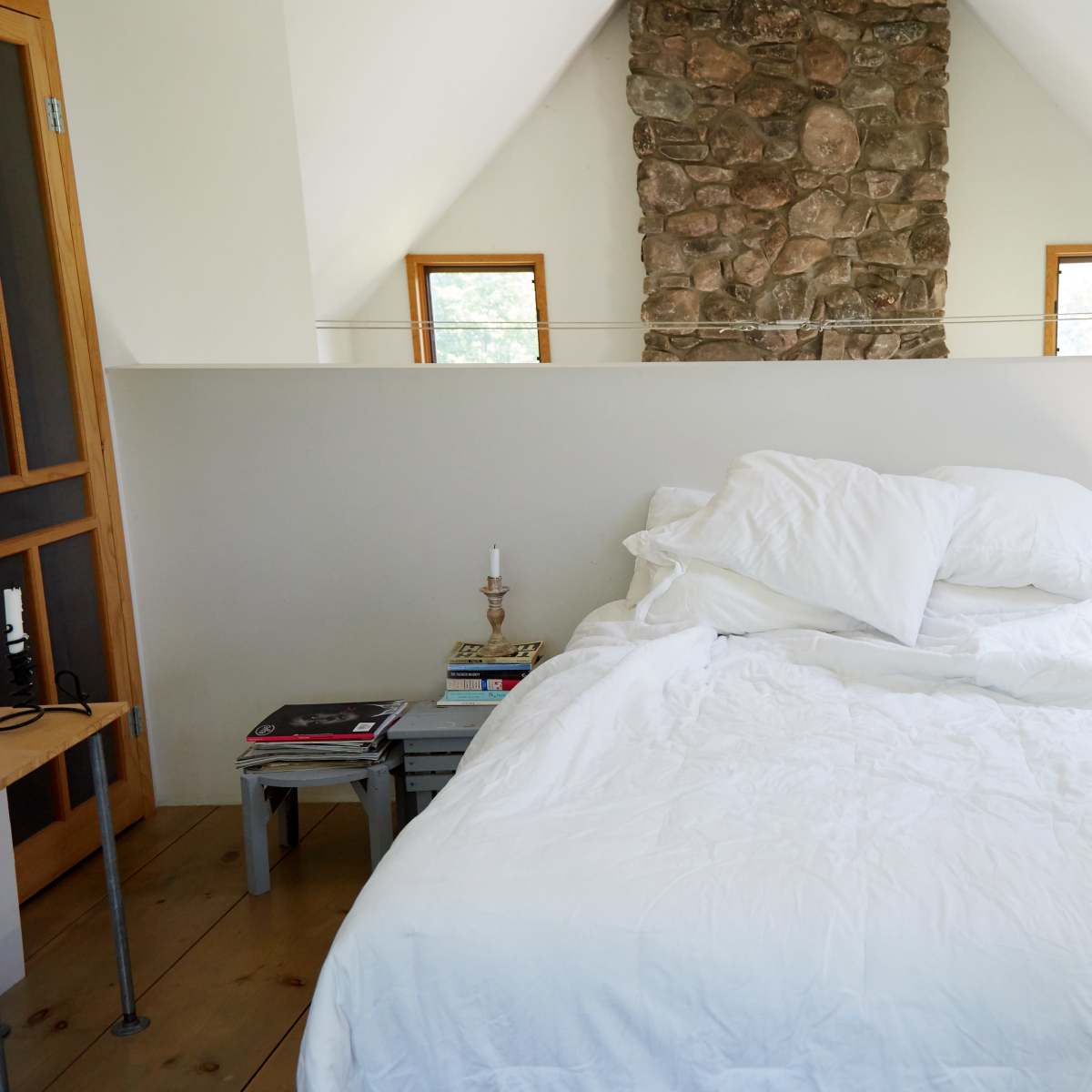 A simple bedroom features a unmade bed with white bedding, a stone wall in the background, and a telephone on a nightstand.