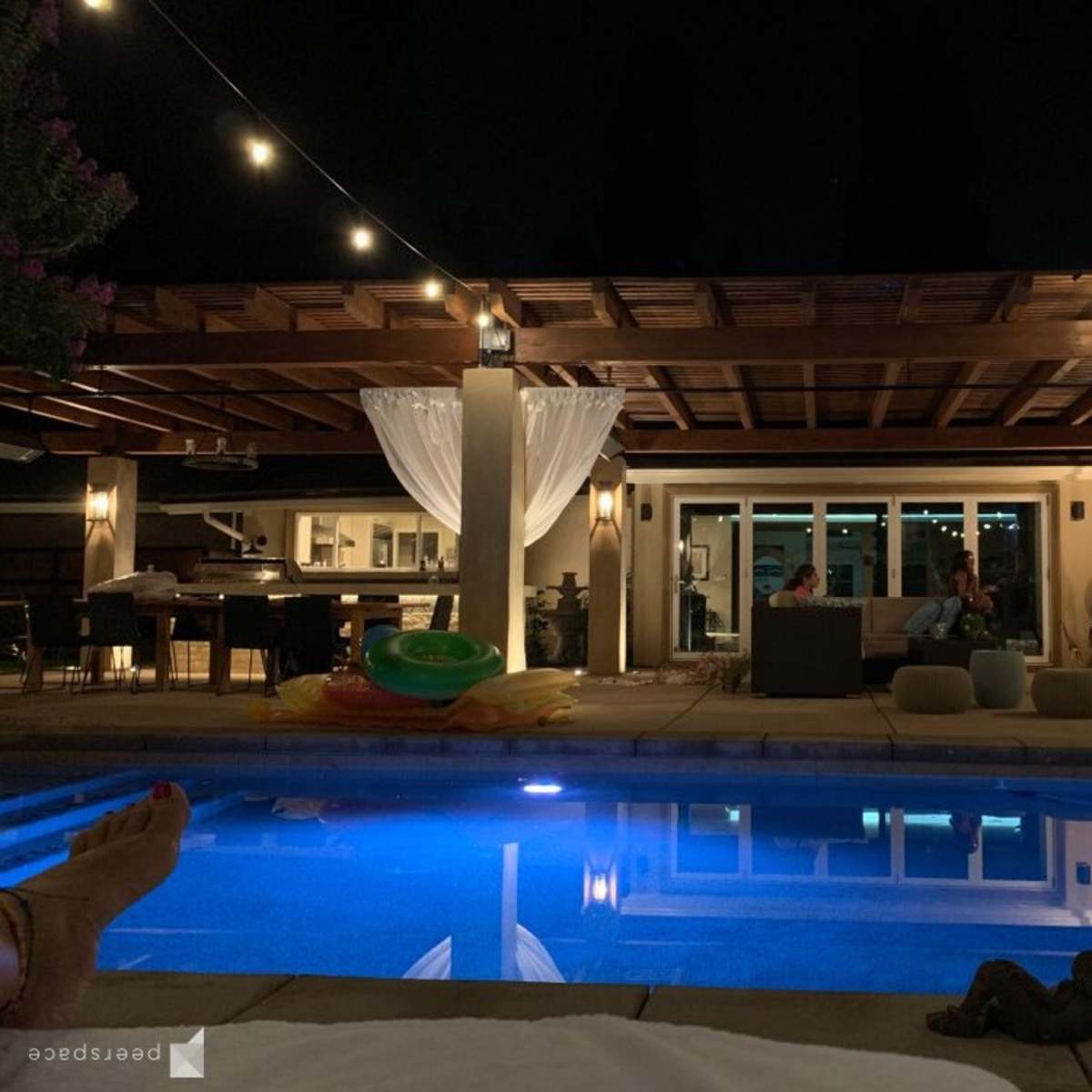 A well-lit outdoor space features a swimming pool reflecting lights, a seating area, and a wooden pergola adorned with string lights at night.