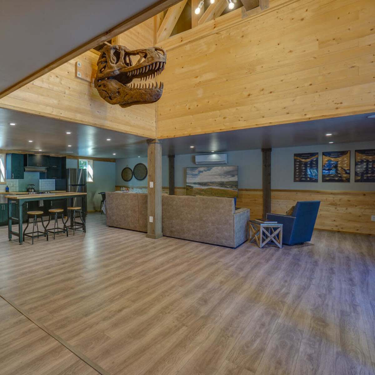 A spacious interior with wooden walls and flooring, featuring a staircase leading to an upper level, a kitchen area in the background, and a seating area with a couch and a chair.