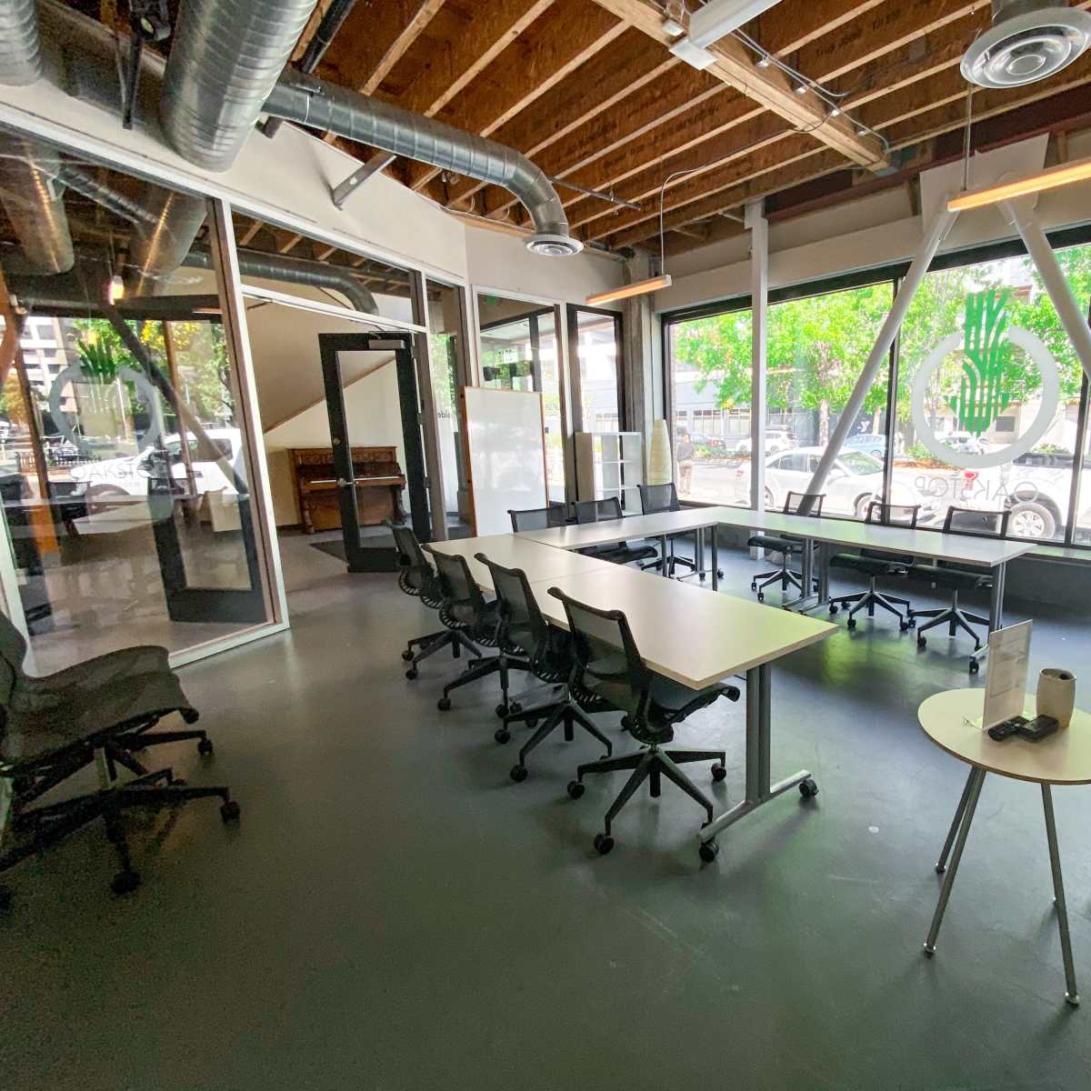 A modern office workspace features several tables and chairs arranged in a bright area with large windows and exposed beams.