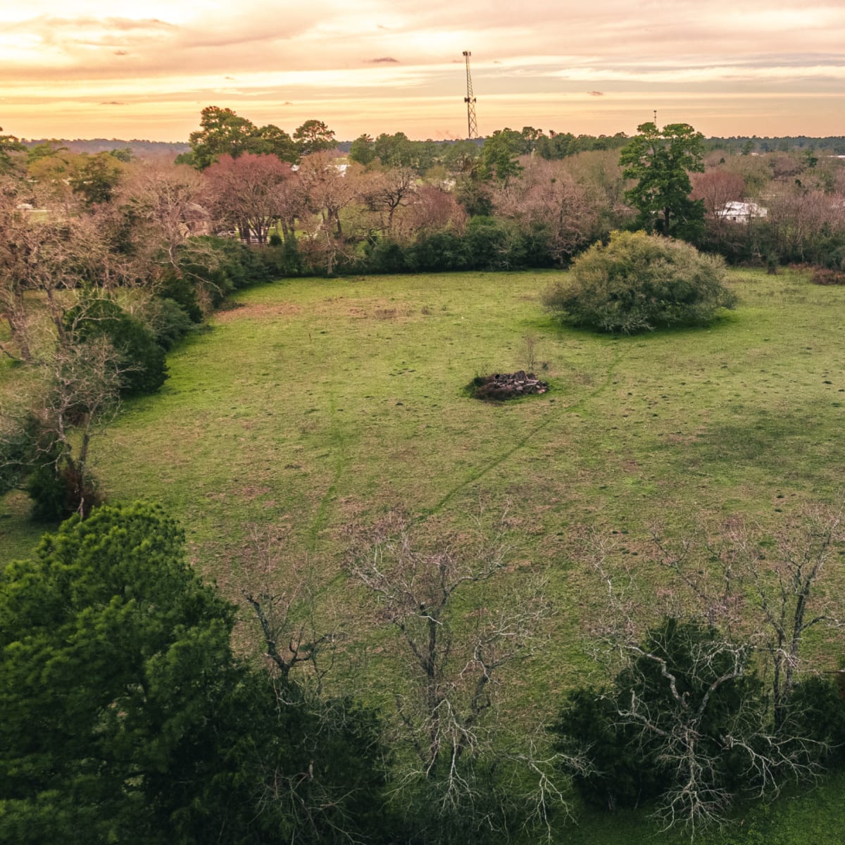 An aerial view of a wide, grassy field surrounded by sparse trees under a cloudy sky.