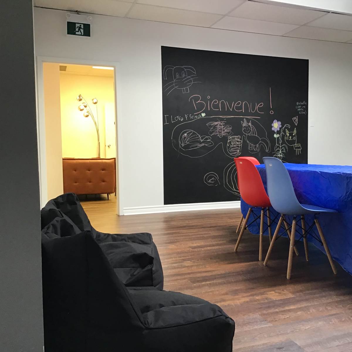 A room with a large blue table surrounded by colorful chairs, a chalkboard wall with drawings and text, and a cozy seating area in the background.