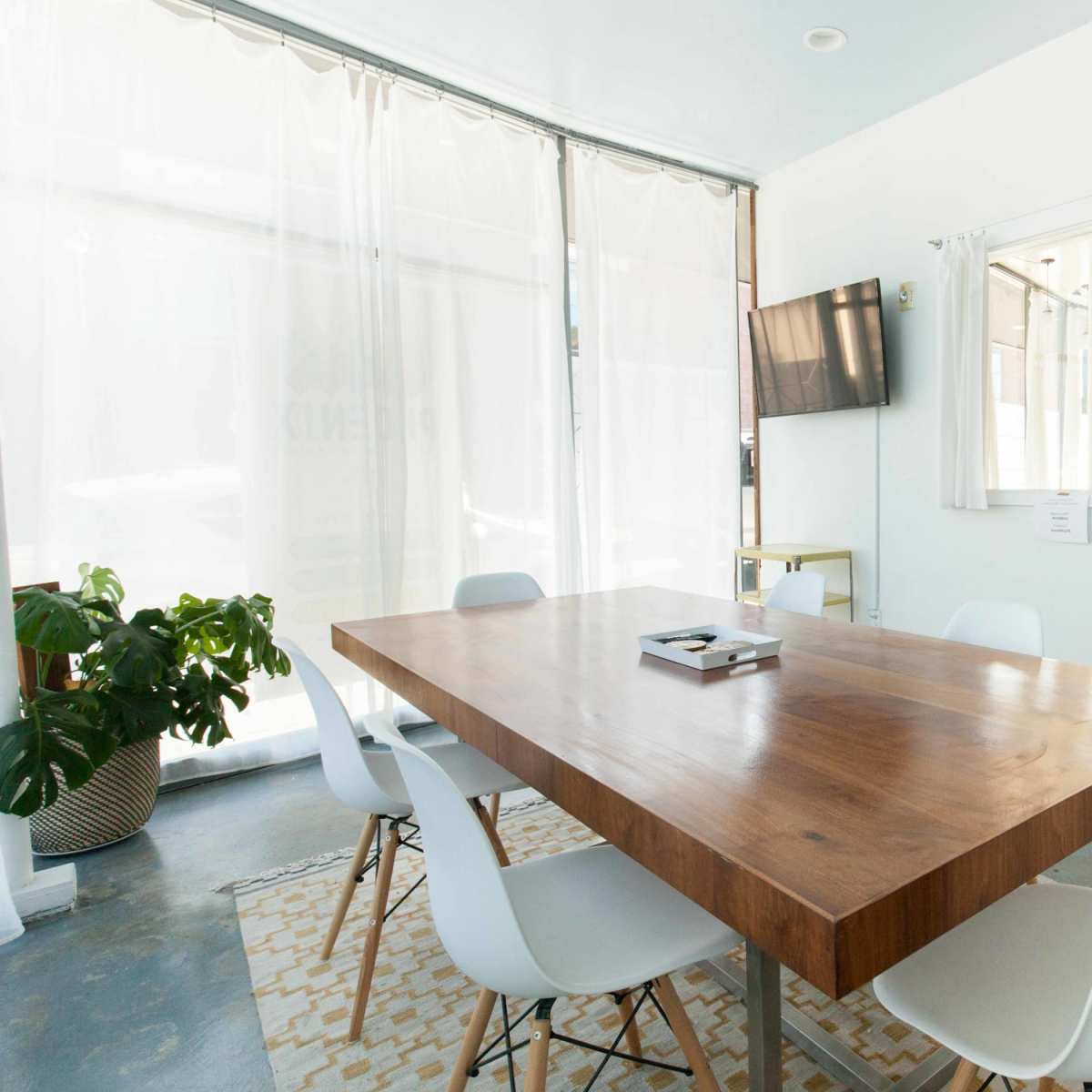 A modern meeting room features a large wooden table surrounded by white chairs, with large windows letting in natural light and a TV mounted on the wall.