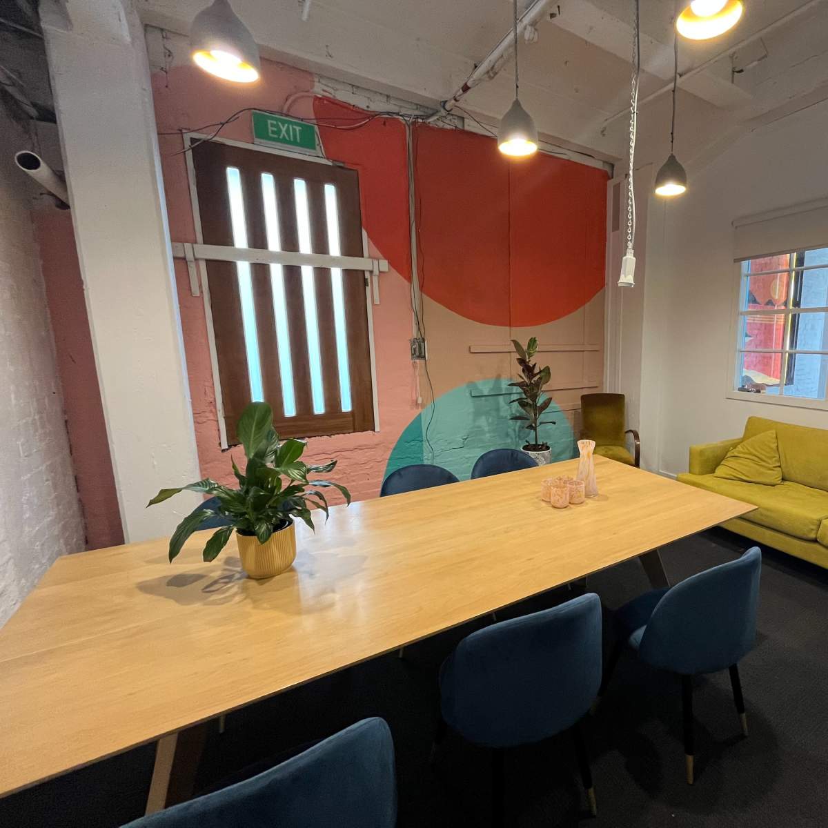 A modern meeting room features a large wooden table, blue chairs, a yellow couch, and colorful wall art with plants decor.