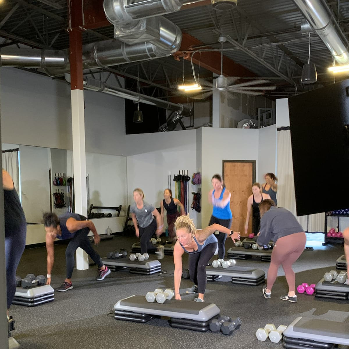 A group of people exercises in a fitness studio while using weights and step platforms.