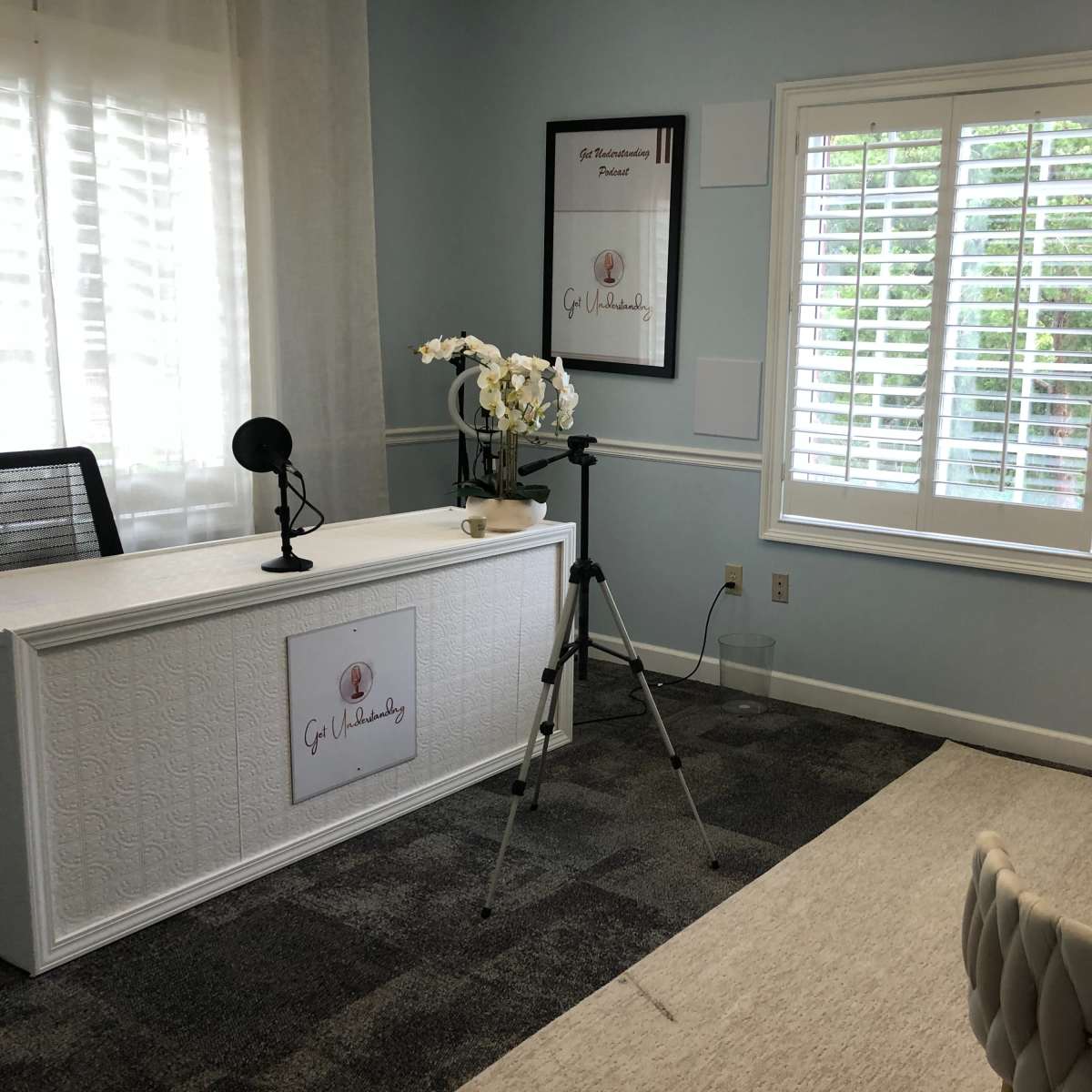 A light blue room with a desk featuring a logo, a microphone setup, a camera on a tripod, and a white chair.
