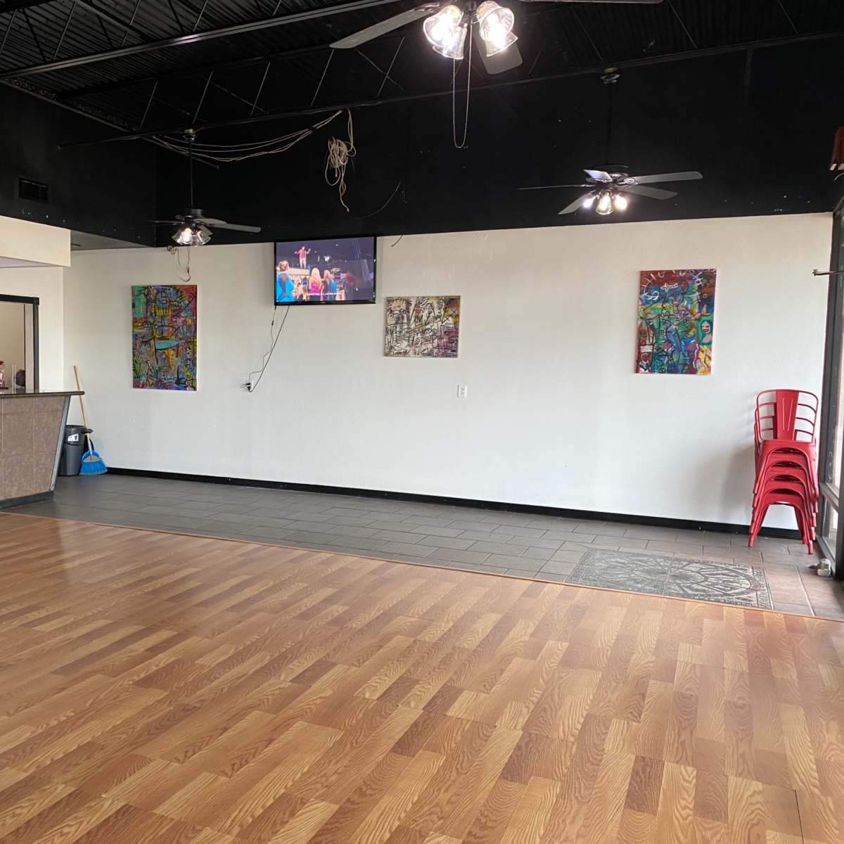 A spacious restaurant interior with a wooden floor, a service counter, a wall-mounted TV, and colorful artwork displayed on the walls.
