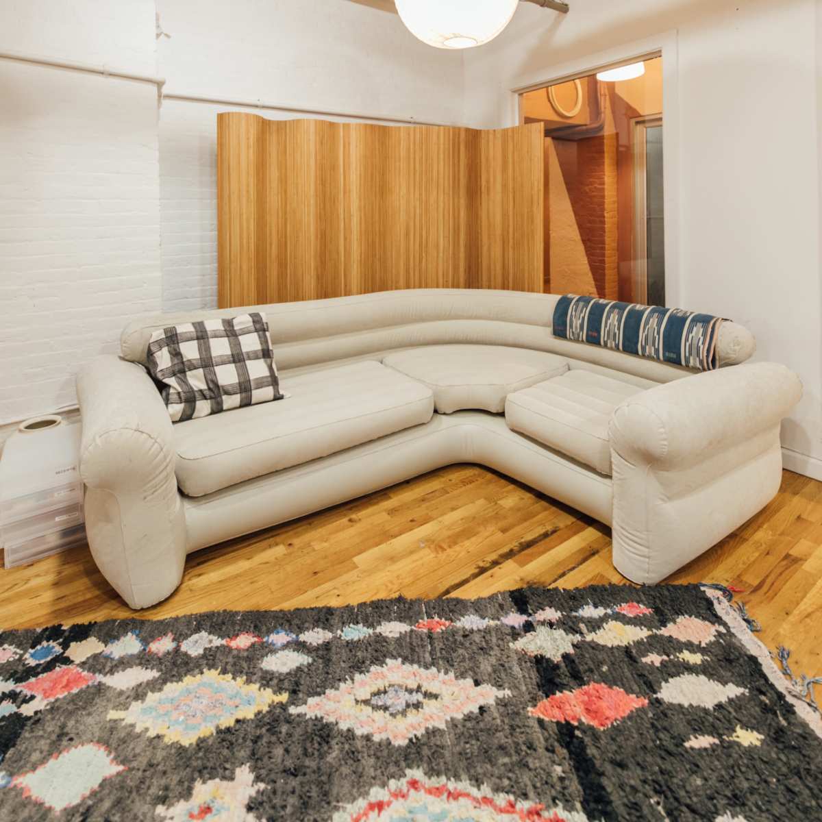A modern, light-colored sectional sofa positioned on a patterned rug in a room with exposed brick walls and overhead lighting.