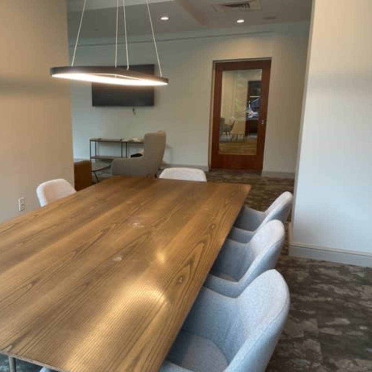 A conference room with a large wooden table surrounded by several light-colored chairs, and a wall-mounted screen in the background.