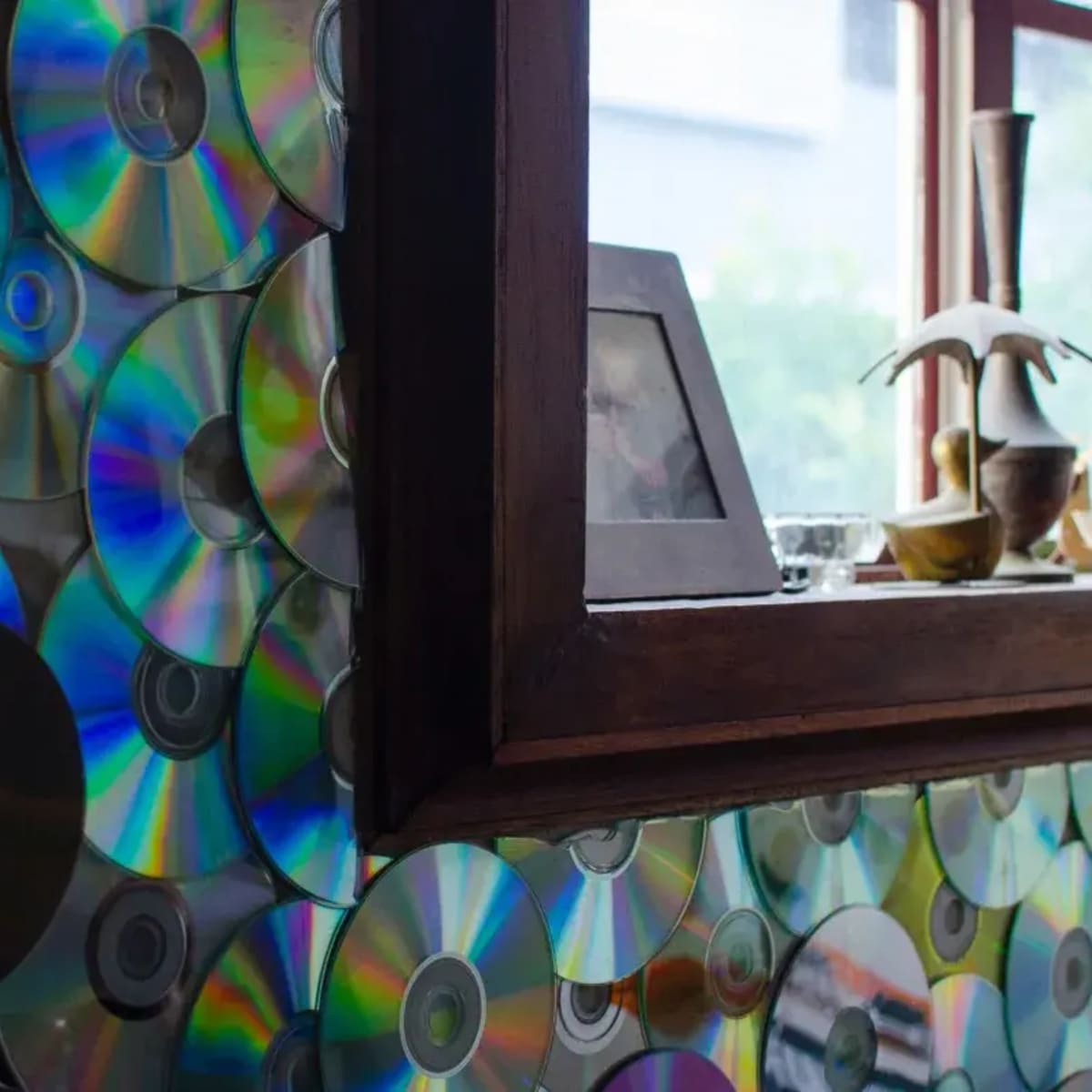 A wall covered with compact discs, reflecting light and arranged around a wooden framed mirror.