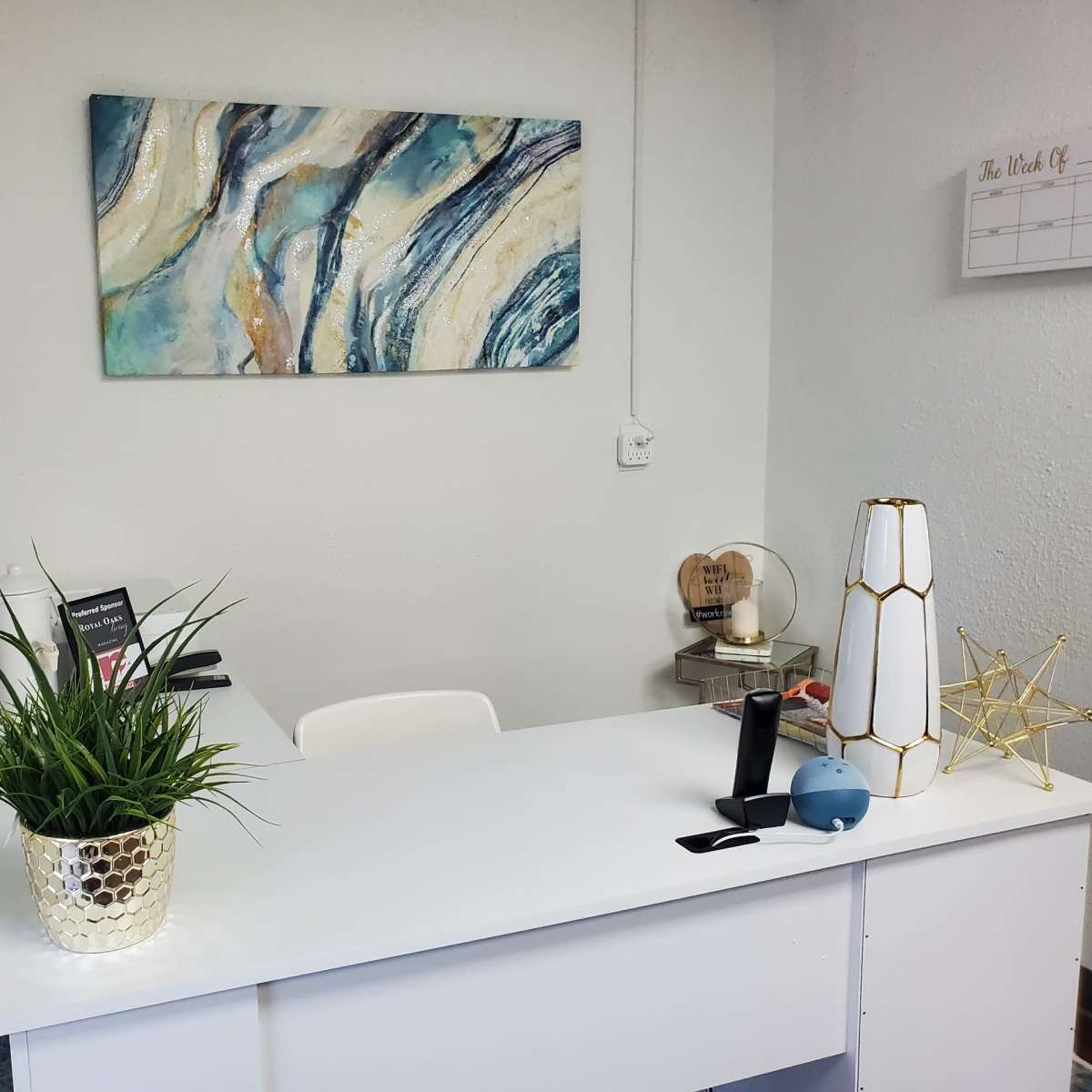 A modern office reception area with a white desk, a decorative abstract painting on the wall, and a potted plant.