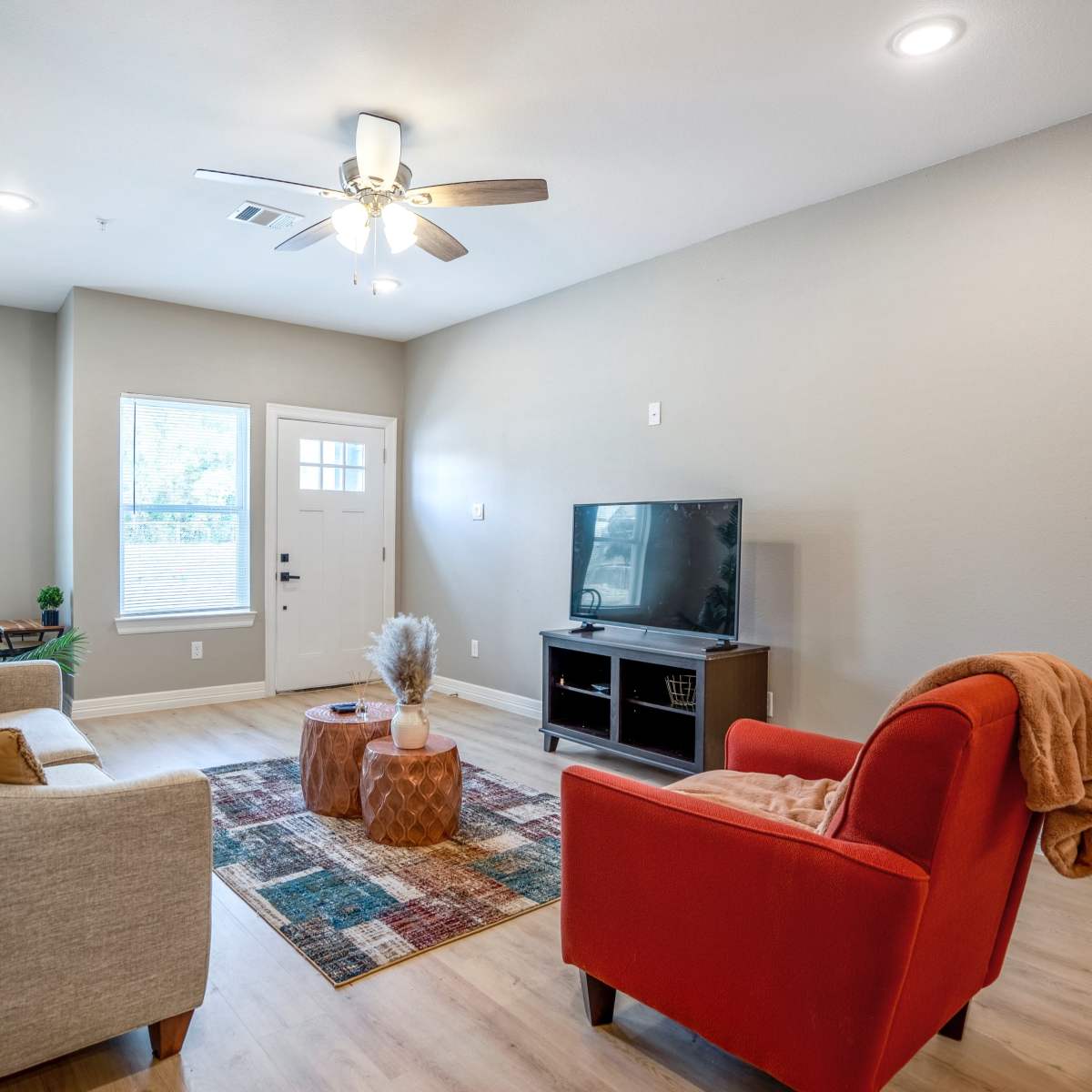The living room features a beige sofa, a red armchair, a wooden TV stand, and a colorful area rug beneath a ceiling fan and natural light from the windows.