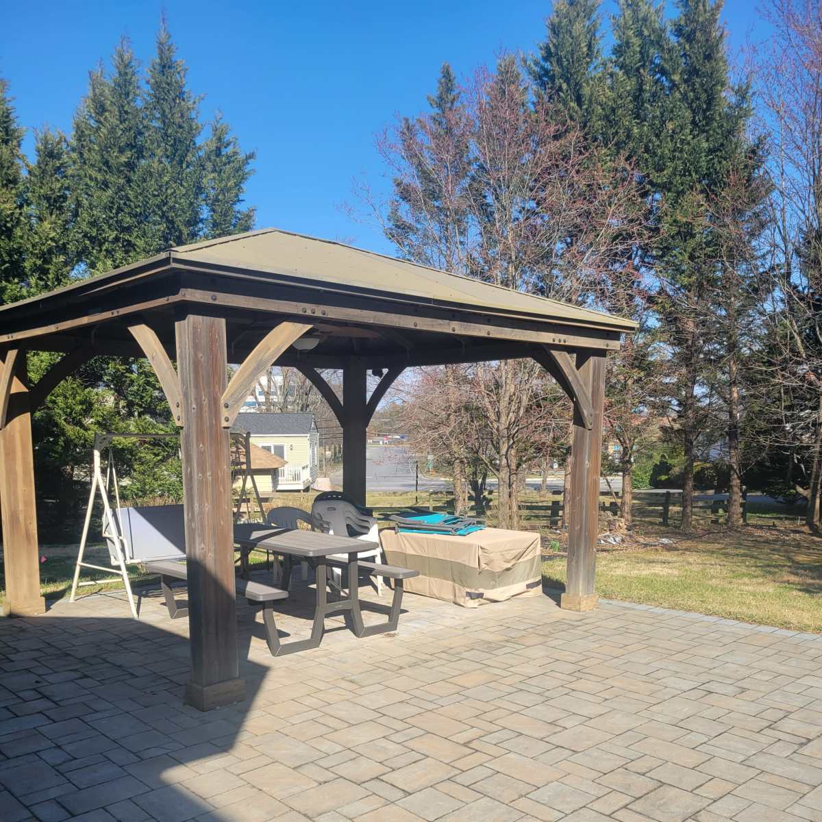 A wooden gazebo with a tan roof overlooks a stone patio area equipped with a table and chairs, alongside a covered hot tub and grassy surroundings.
