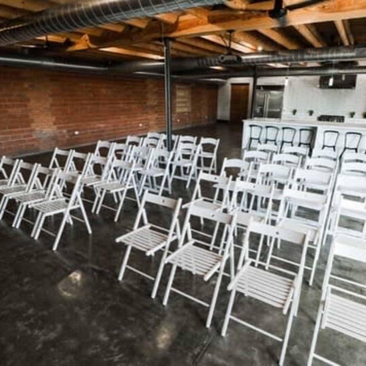 A row of white folding chairs is arranged in a spacious, industrial-style room with exposed brick walls and a concrete floor.