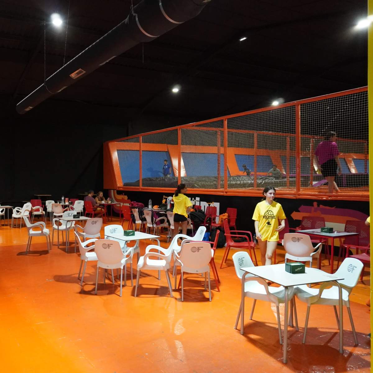 An indoor trampoline park with an adjacent seating area featuring brightly colored chairs and tables, while people can be seen jumping on trampolines behind a safety net.