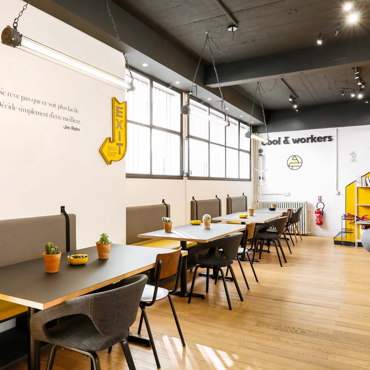 A modern workspace with long tables, potted plants, and a large exit sign on the wall.