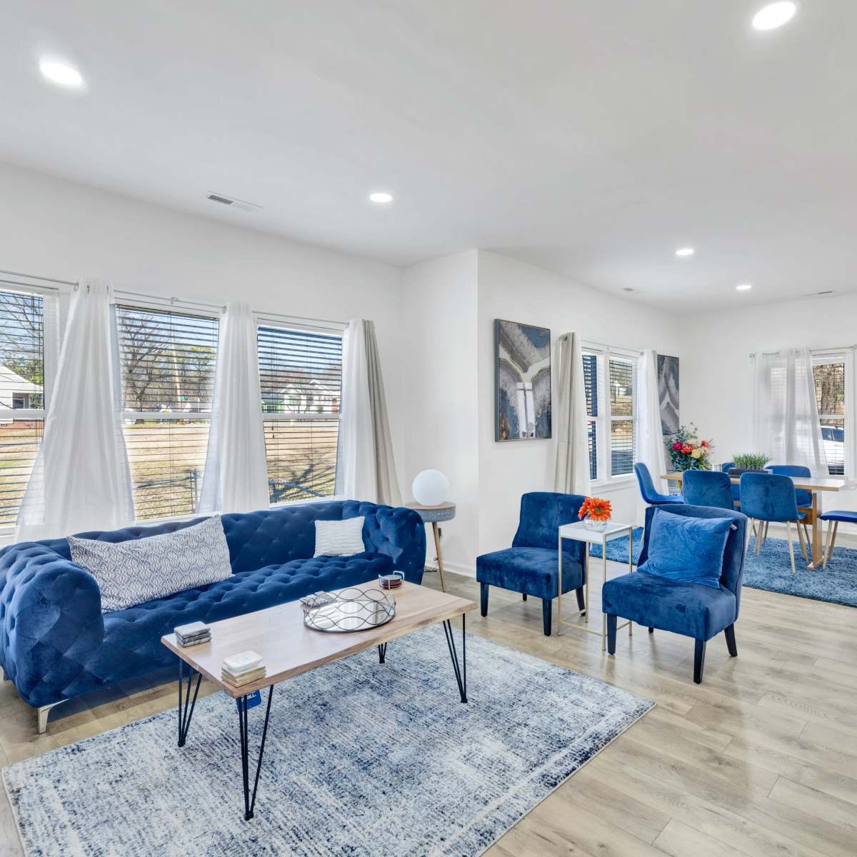 The living room features a blue sofa and chairs arranged around a coffee table, with large windows letting in natural light.