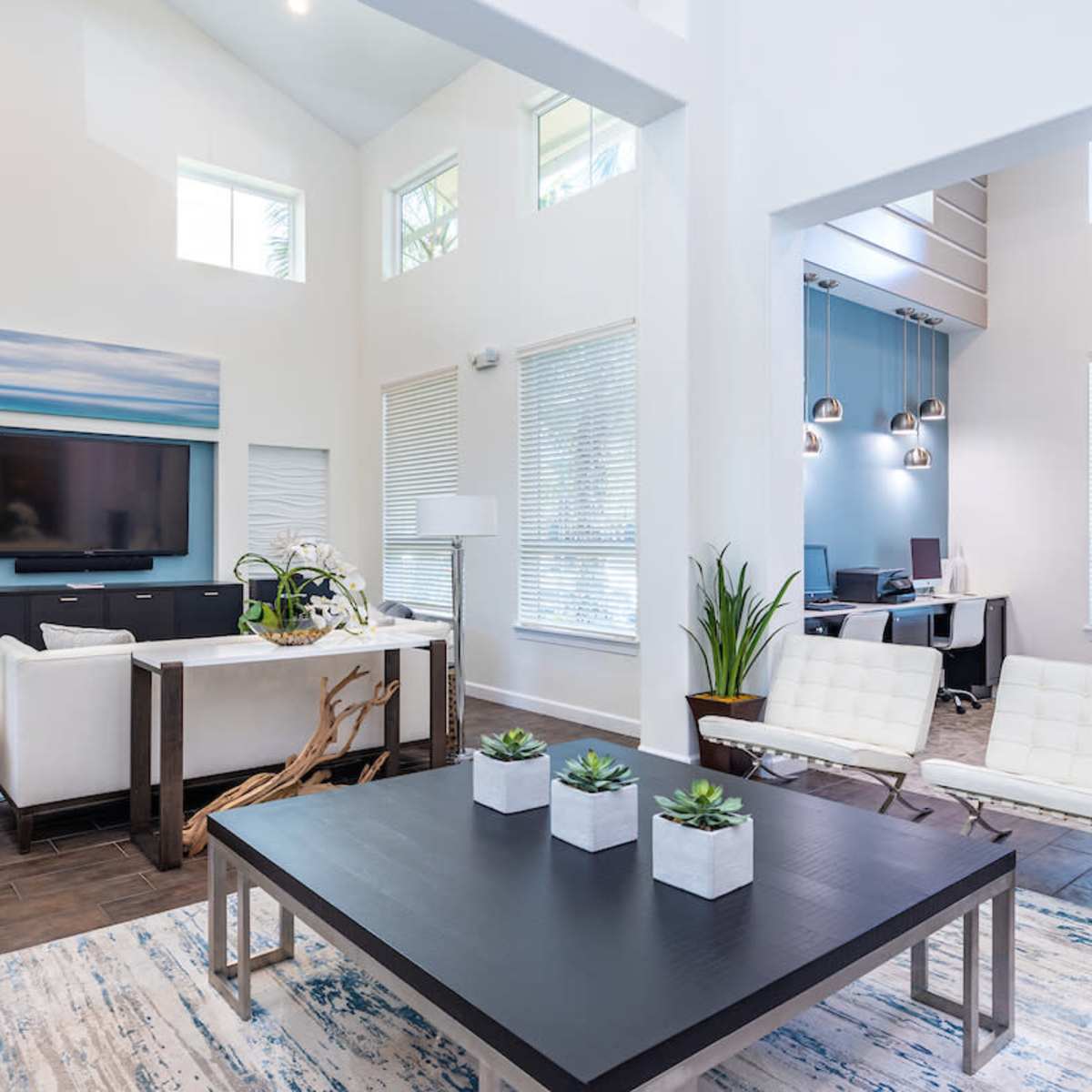 A modern living room with a white couch, a large coffee table, a television mounted on the wall, and a workspace area visible through an open archway.