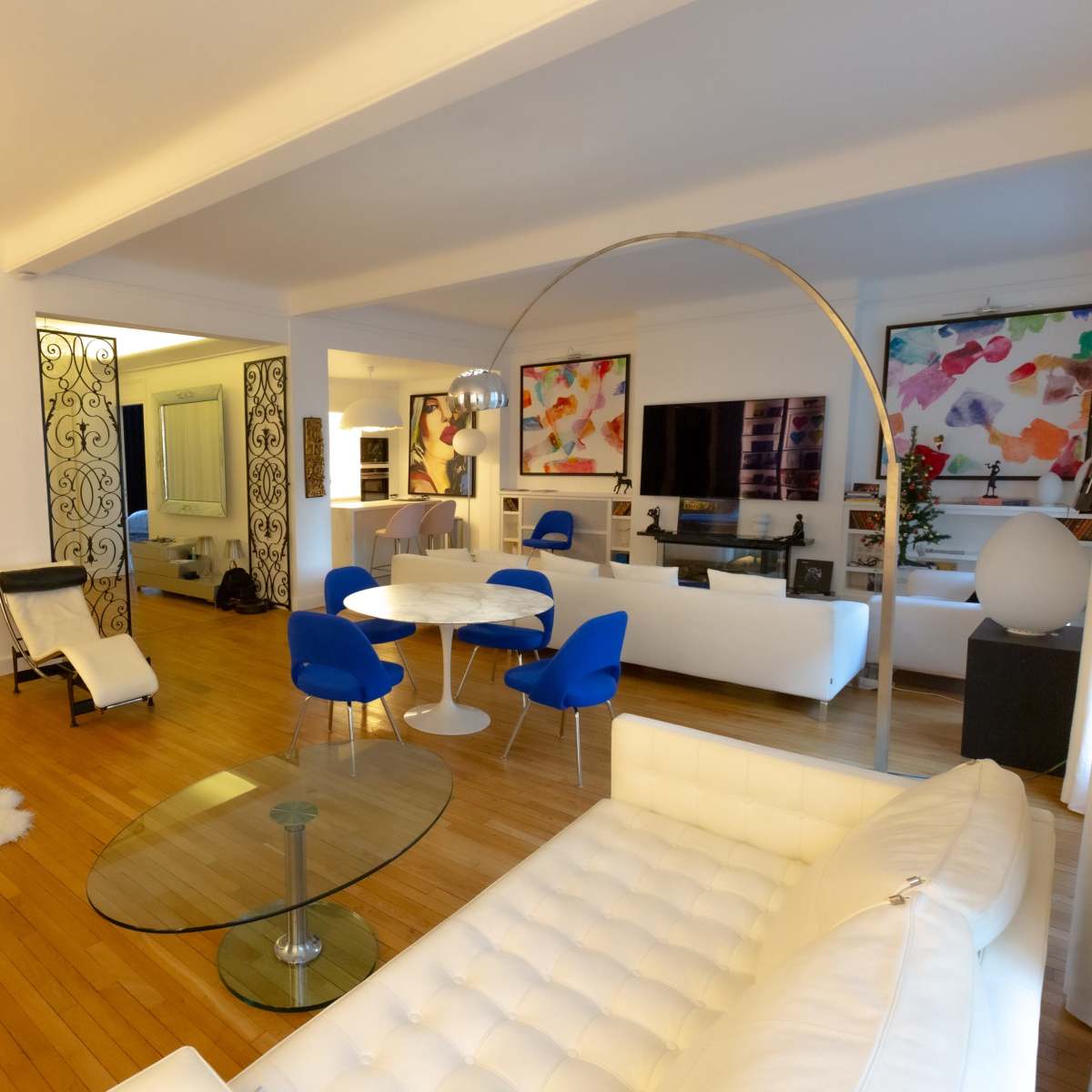 A modern living room featuring a white leather sectional sofa, a circular dining table with blue chairs, and large abstract art pieces on the walls.