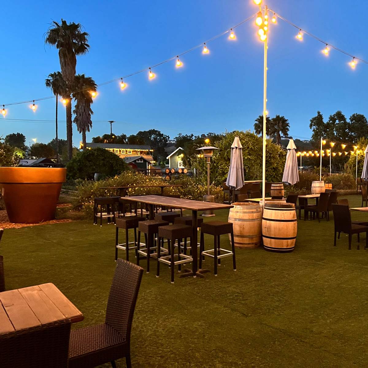 The outdoor dining area features wooden tables, high stools, and string lights, surrounded by potted palms and umbrellas at dusk.