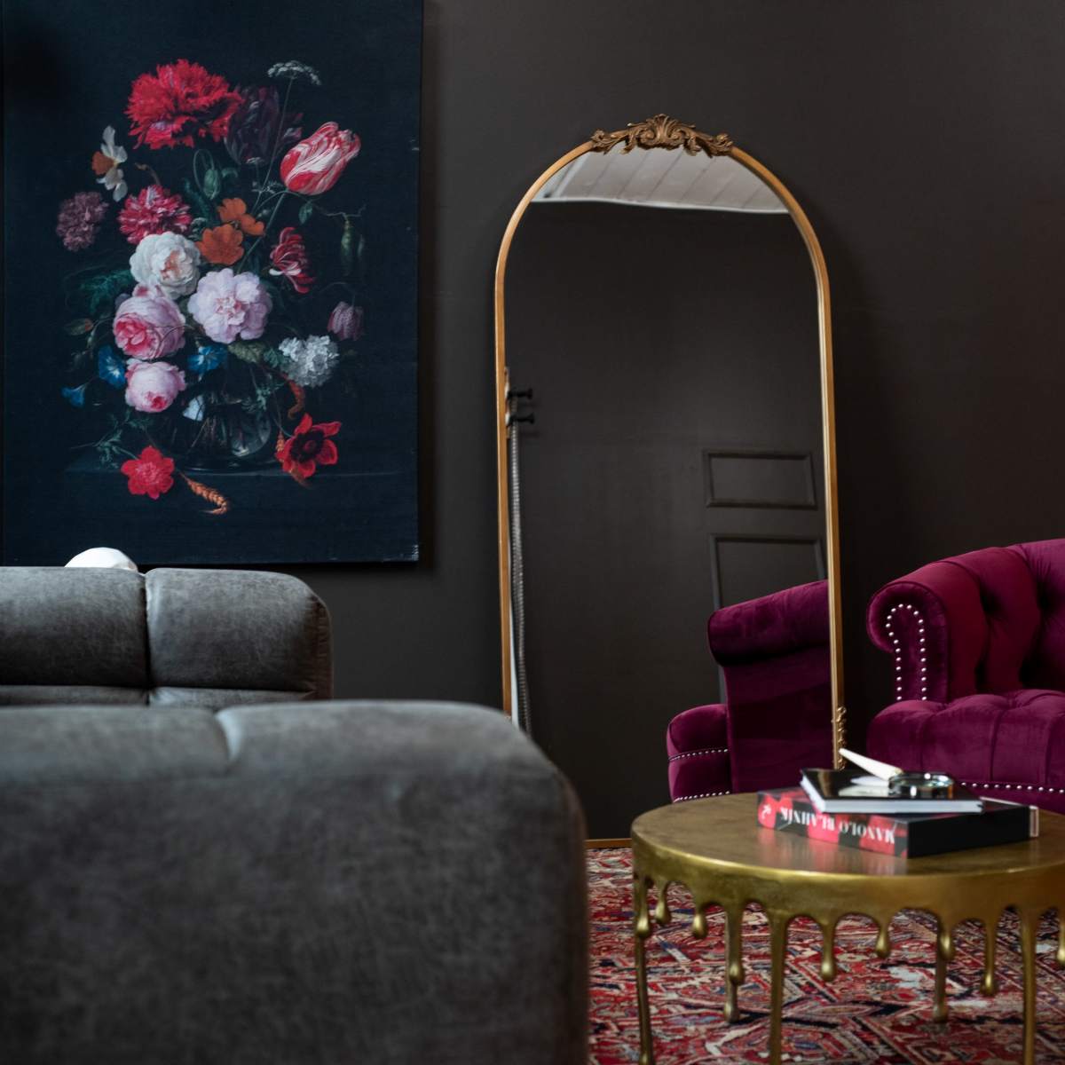 A modern living space features a grey sofa, a purple tufted armchair, a circular gold coffee table with books, and a large floral painting on the wall opposite an ornate mirror.