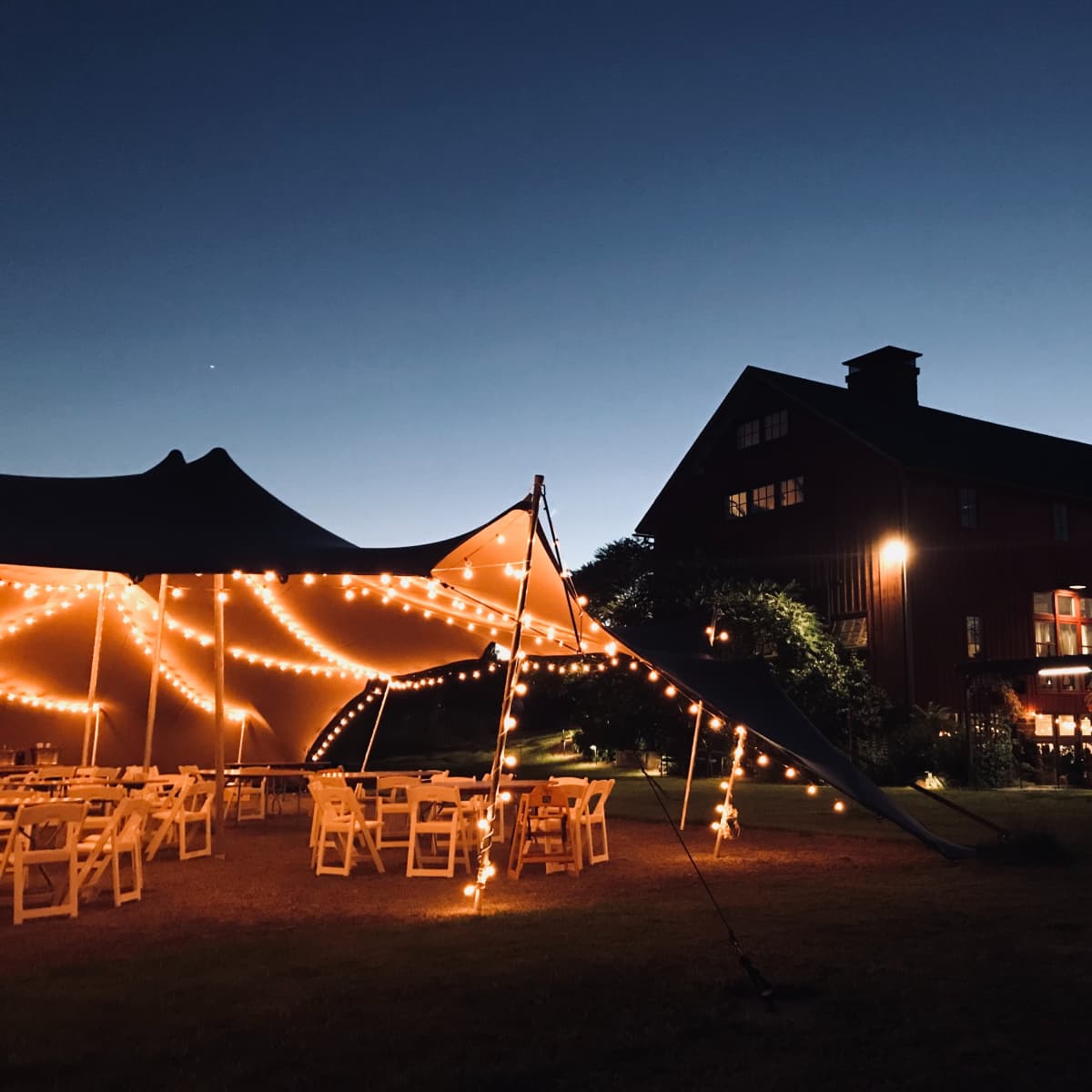 A large red barn sits beside a lighted tent with tables, illuminated against a twilight sky.