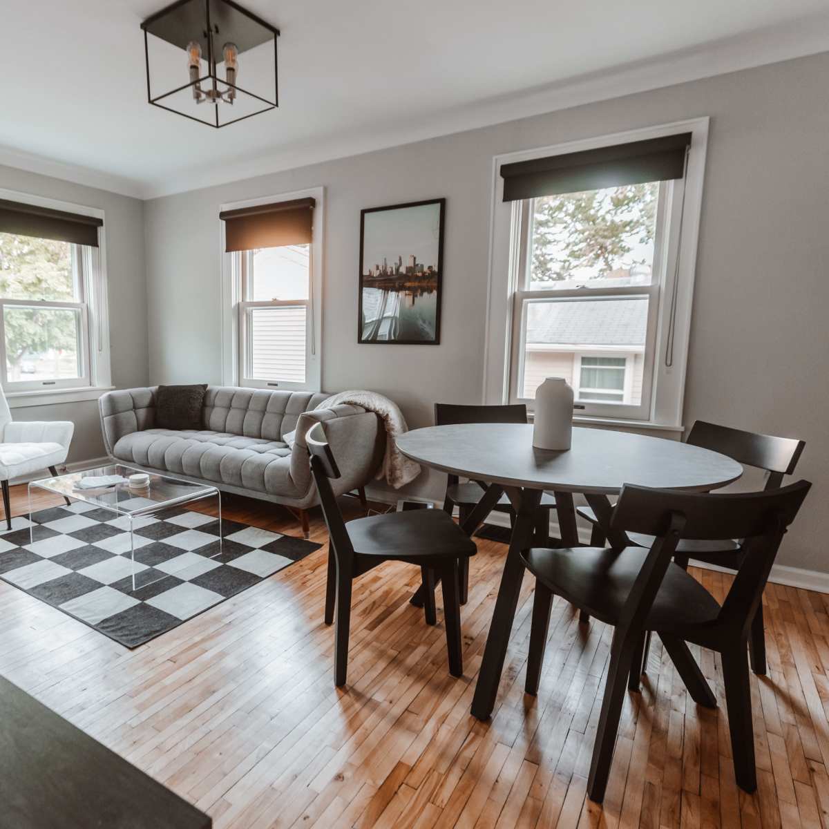 A modern living space featuring a gray sofa, a round black dining table with chairs, and a checkered rug on hardwood flooring.