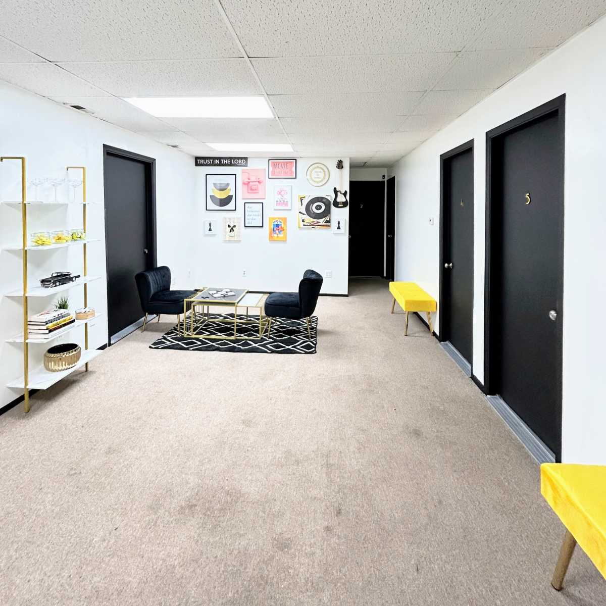 A hallway with four black doors, a coffee table with two black chairs, and art pieces on the wall, all set on a carpeted floor.