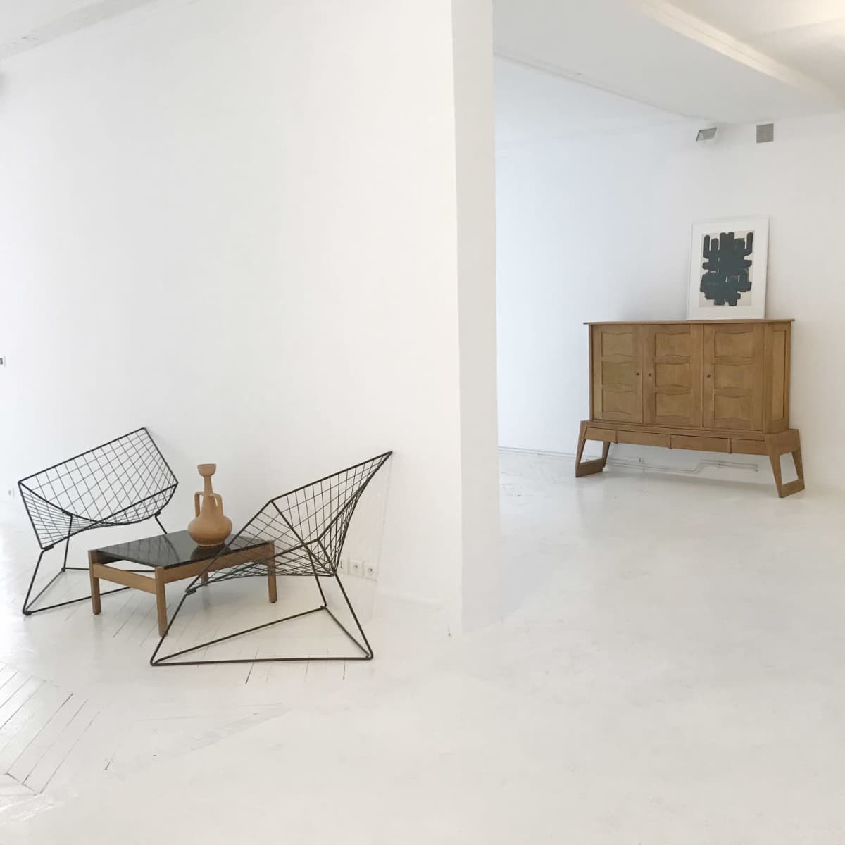 An open, white-walled room features two wire chairs, a wooden side table, and a wooden cabinet against the wall.