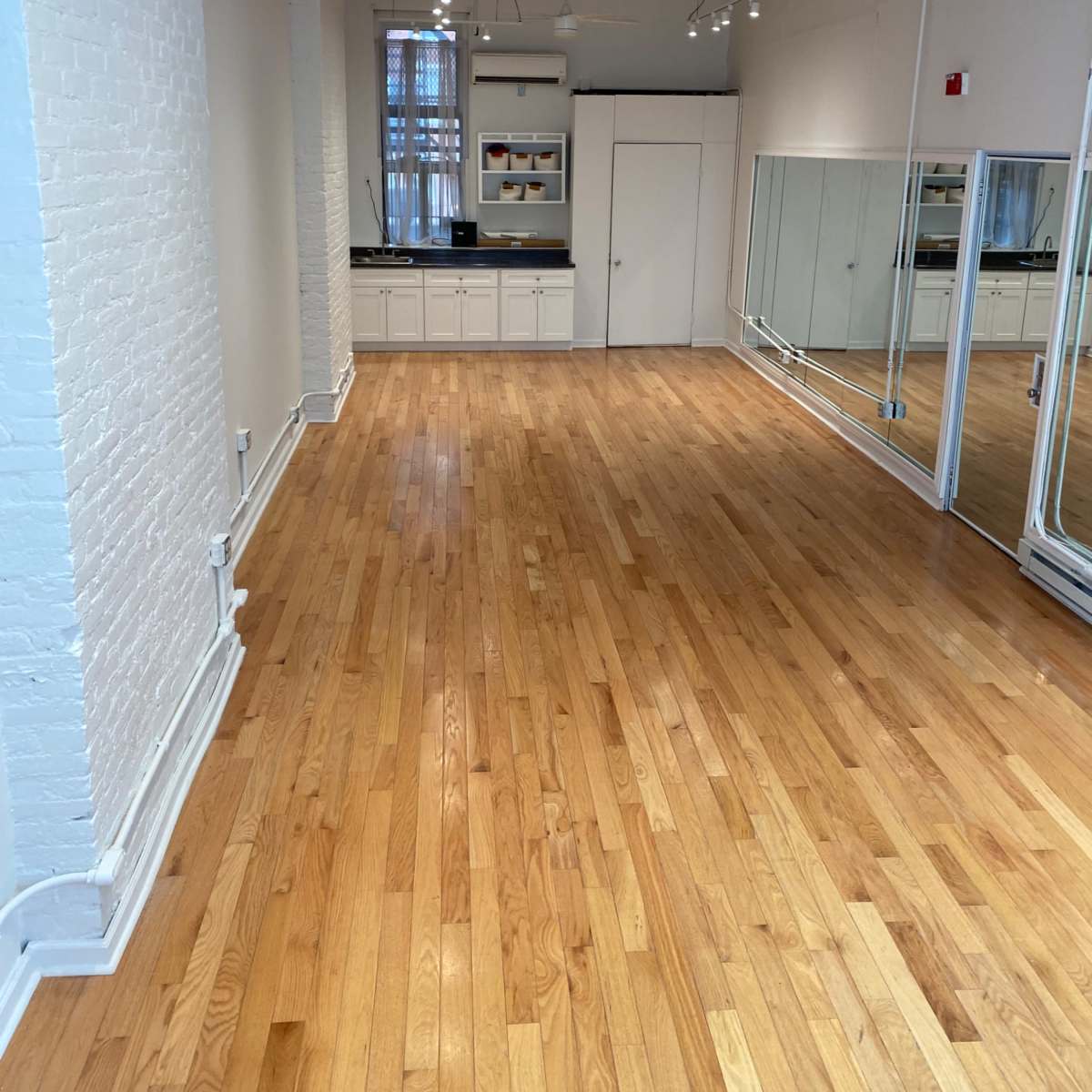 An empty room with polished wooden flooring, white brick walls, and a mirrored wall at one end, along with a kitchenette area featuring cabinets.