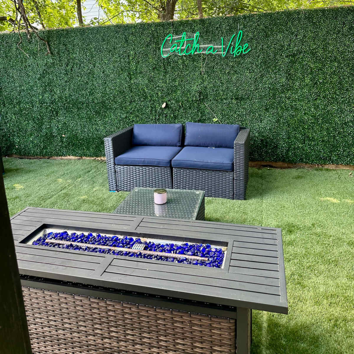 A cozy outdoor space features a rattan couch with navy cushions beside a fire pit adorned with blue glass stones, under a green hedge and a neon sign that reads "Catch a Vibe."