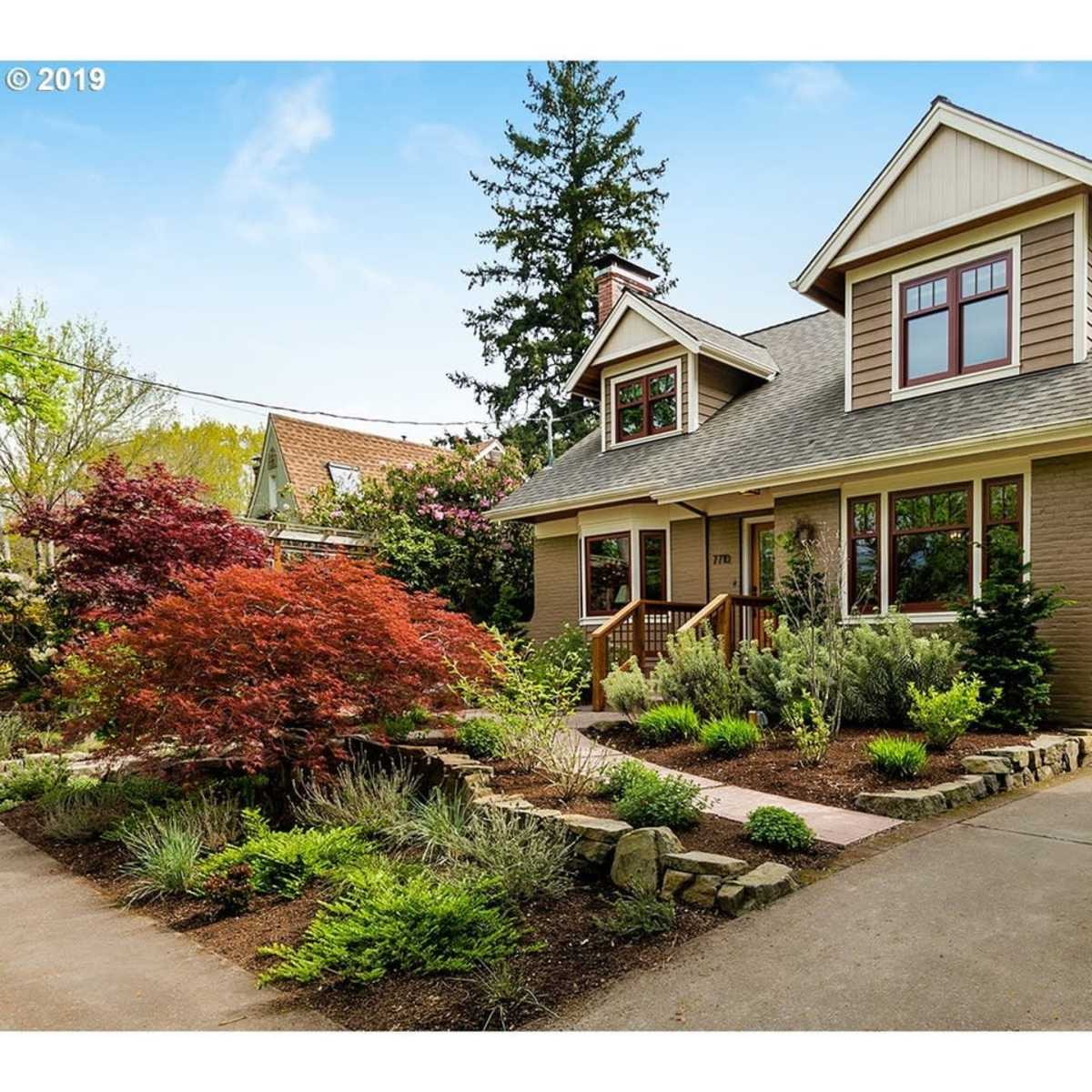A two-story, craftsman-style house with a landscaped front yard features a mix of shrubs and small trees.