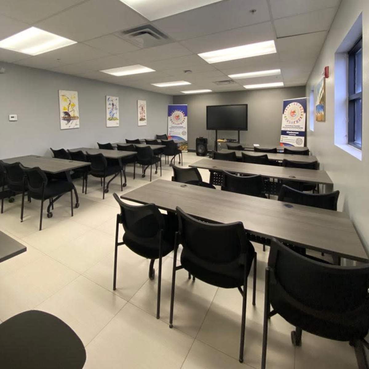 A classroom or meeting room with multiple rows of black tables and chairs, a large screen at the front, and posters on the walls.