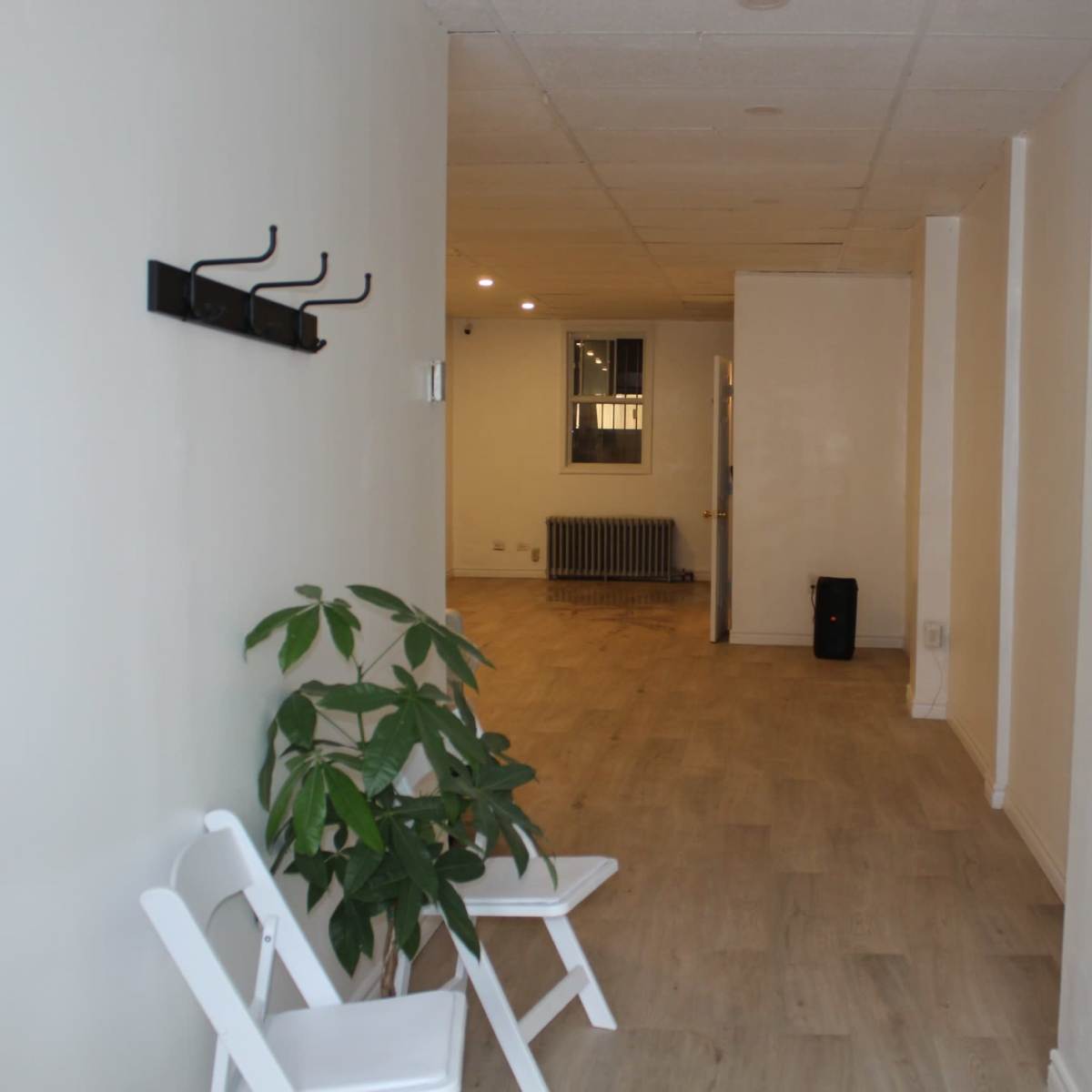 A long, empty hallway with light-colored walls, wooden flooring, a potted plant, and some folding chairs positioned against one side.