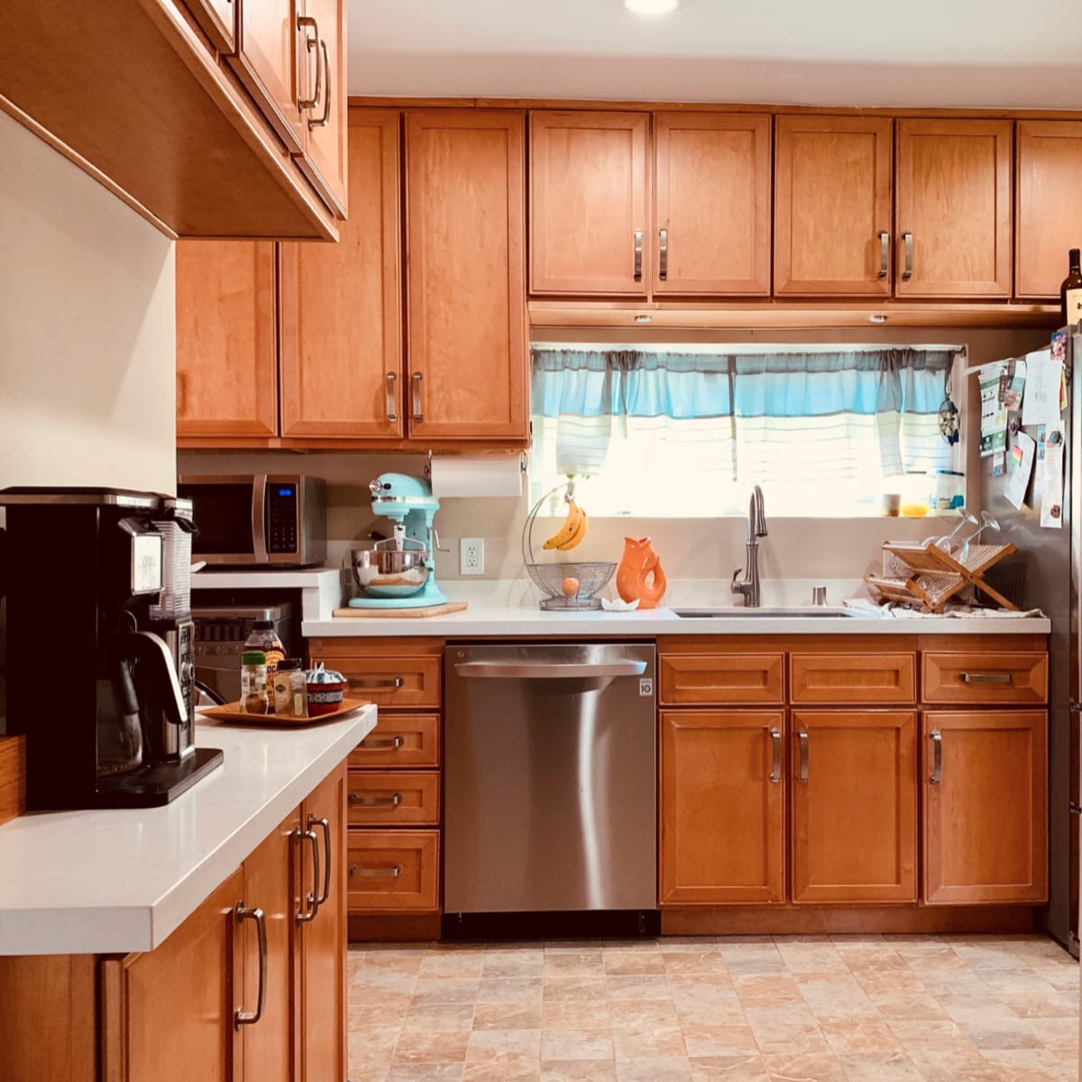 A kitchen features wooden cabinets, a stainless steel dishwasher, and a window above the sink with light blue curtains.