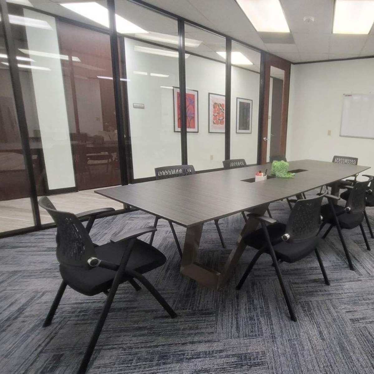 A modern conference room features a long table surrounded by black chairs, with glass walls and a whiteboard in the background.