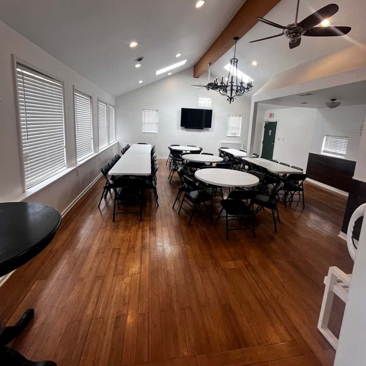 A spacious room with wooden flooring, featuring white walls, multiple round tables with black chairs, and a ceiling fan.