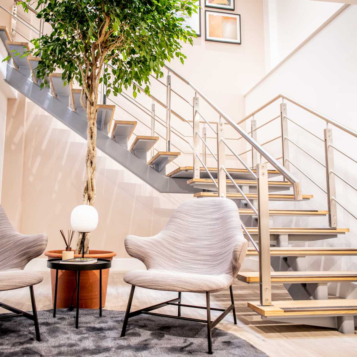 A pair of modern chairs sits beside a small table with a lamp and greenery, next to a staircase with wooden steps and a metal railing.