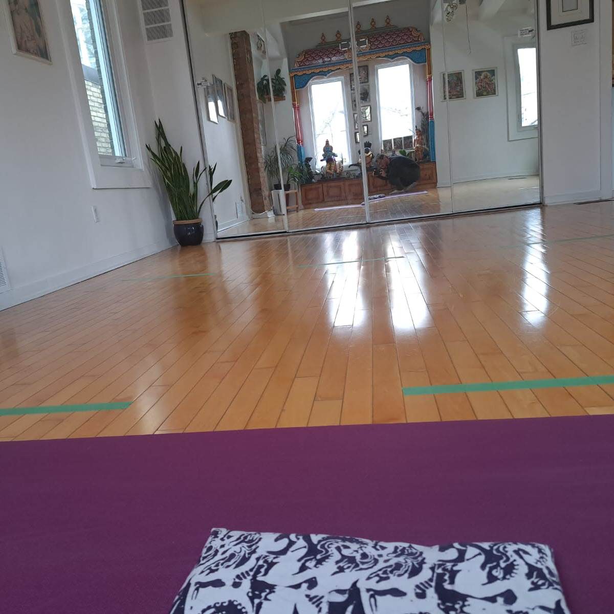 A yoga mat is placed on a polished wooden floor in a spacious room with large mirrors and plants.