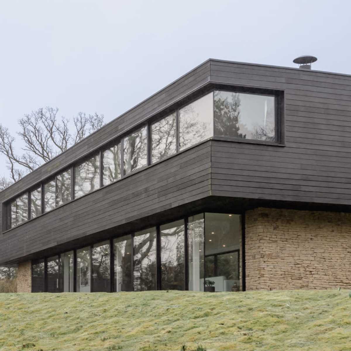A modern two-story house with a combination of stone and black wood siding is situated on a gently sloped grassy area.