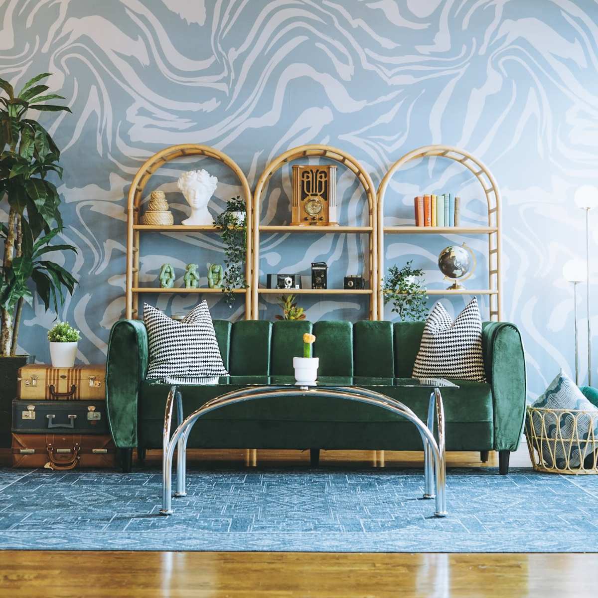 A green velvet sofa is positioned in front of a patterned gray wall, accompanied by a glass coffee table and a stylish shelving unit filled with decorative items and plants.