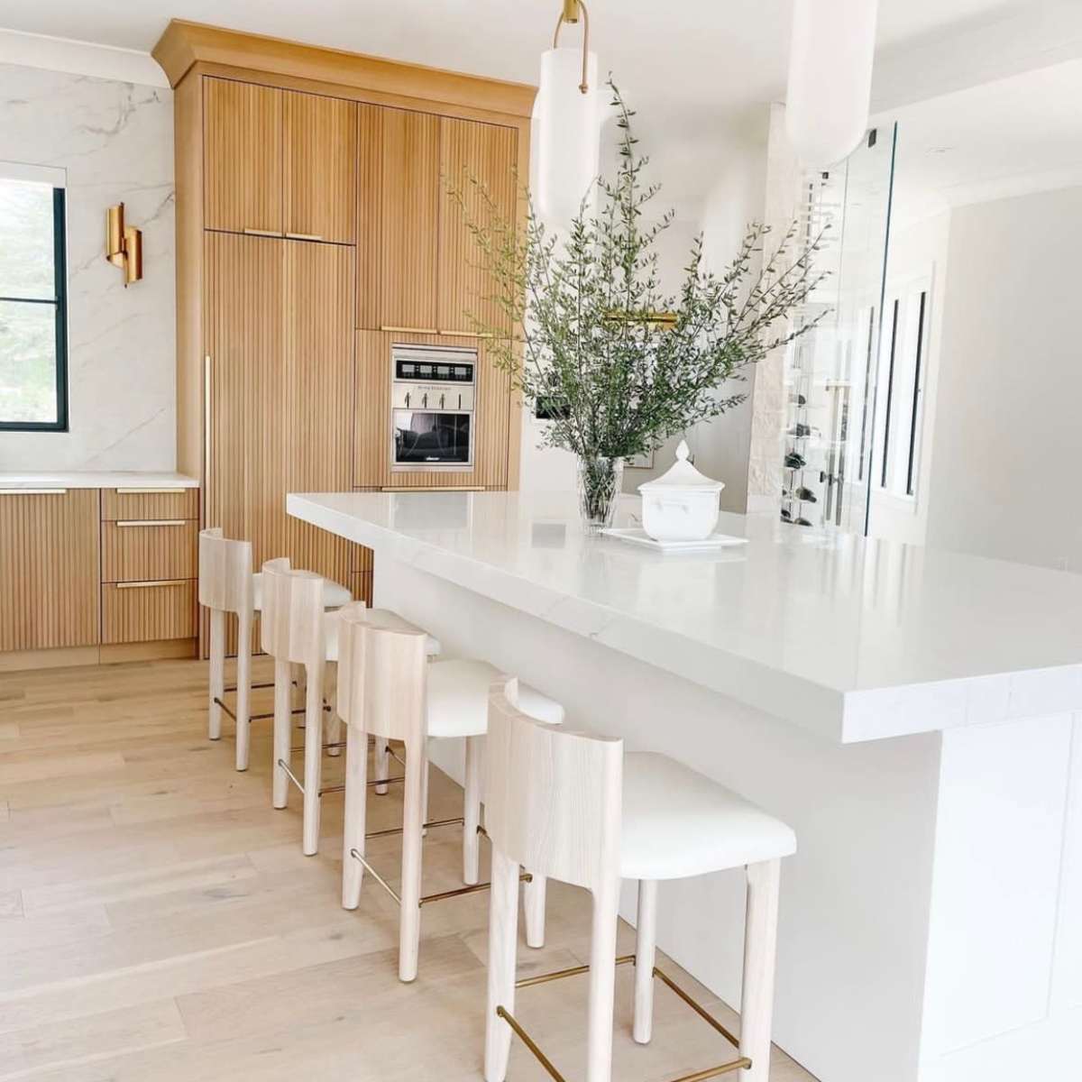 A modern kitchen features a white island with seating and bamboo cabinetry, accented by elegant light fixtures and greenery in a vase.