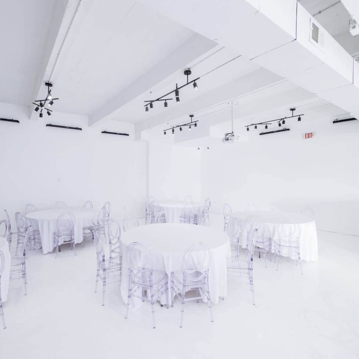 A spacious, minimally decorated white room with several round tables covered in white tablecloths and transparent chairs.