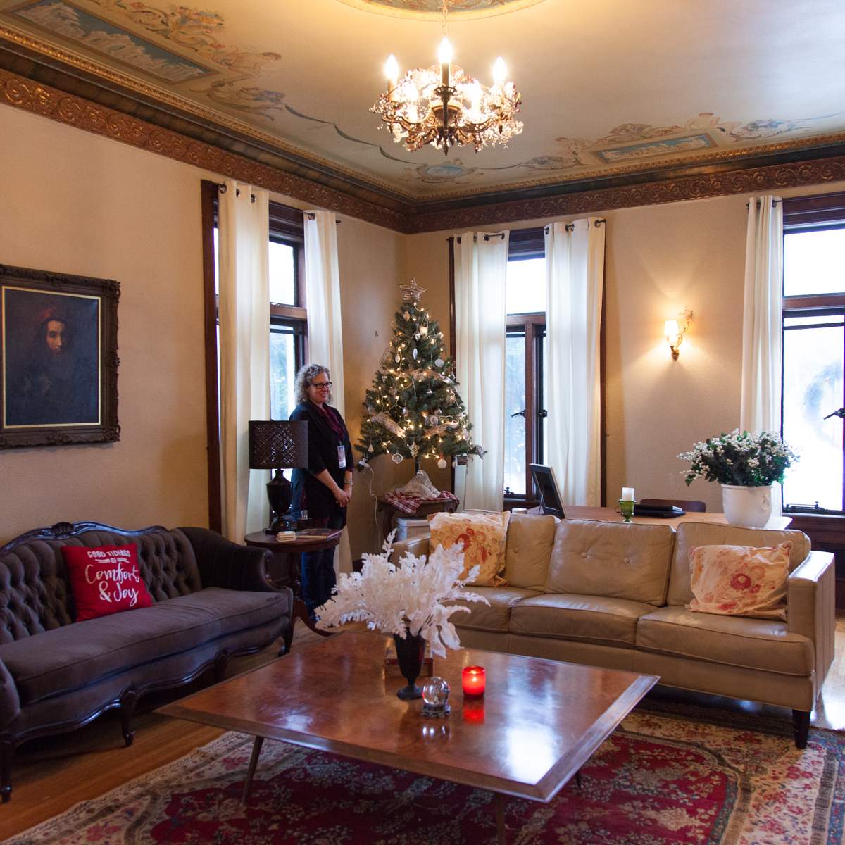 A cozy living room features a decorated Christmas tree, elegant furniture, and a large window adorned with sheer curtains.