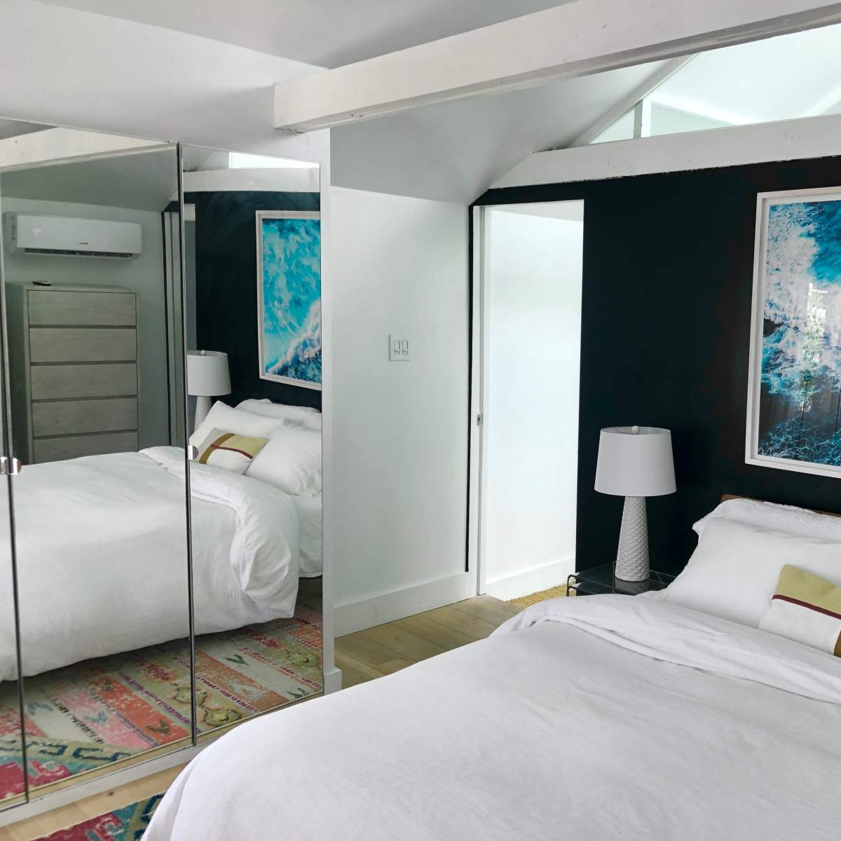 A bedroom with a white bed, a large ocean-themed artwork on the wall, and a mirrored wardrobe reflecting the room's interior.