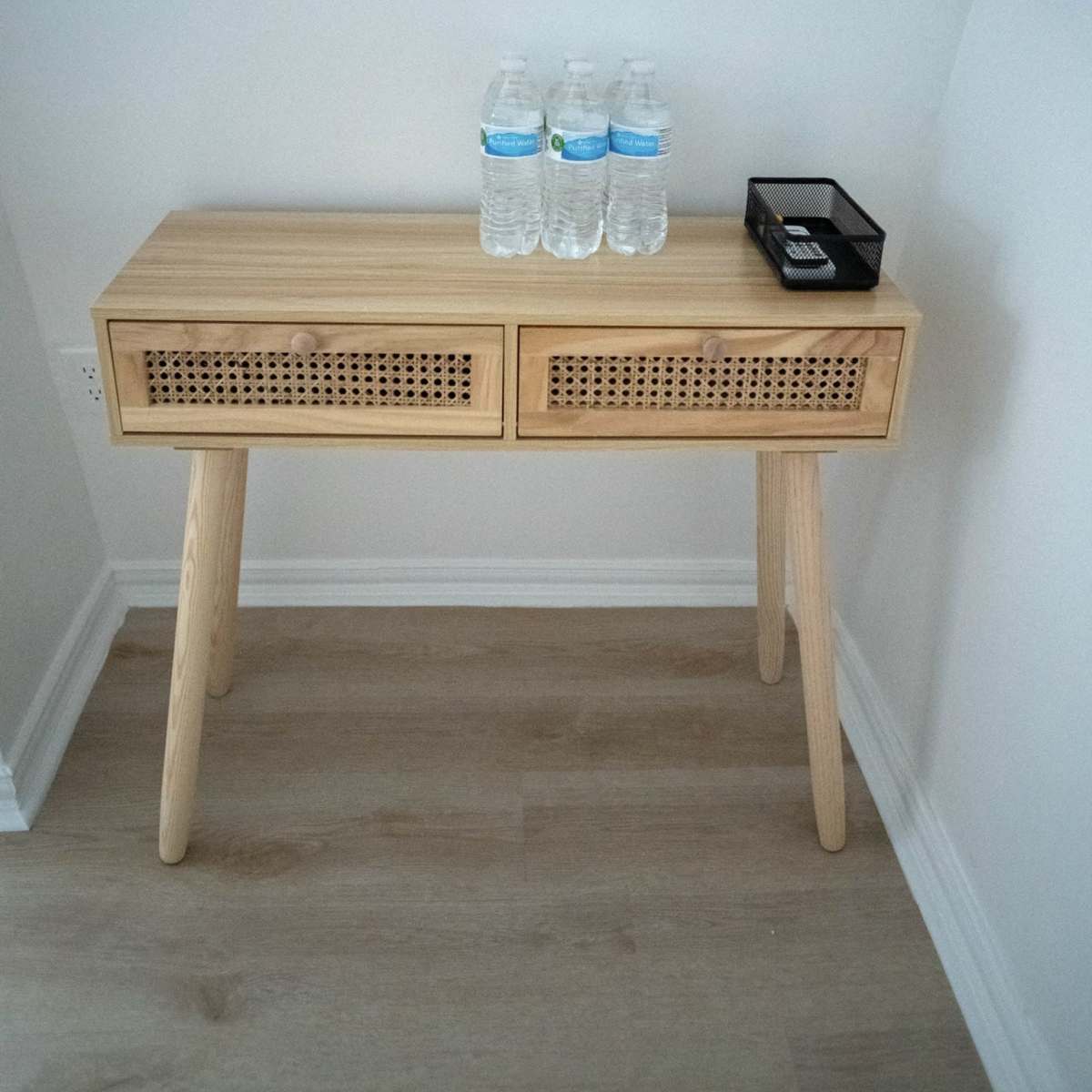 A wooden console table with two drawers stands against a wall, holding three bottles of water and a small black container.