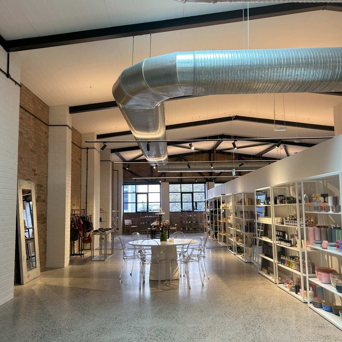 A spacious interior of a contemporary retail store with a large dining table, clear chairs, and shelves filled with various products under industrial-style ductwork.